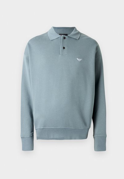Ljusblå tröja, långärmad, treknappskrage, ribbade ärmslut, mjukt material, har en liten broderad logotyp på vänster bröst.