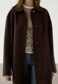 Brauner Wollmantel mit lockerer Passform, ausgestattet mit einem Kragen, sechs Knöpfen und großen Seitentaschen. Tragend über einem hellbeigen Oberteil und Jeans.