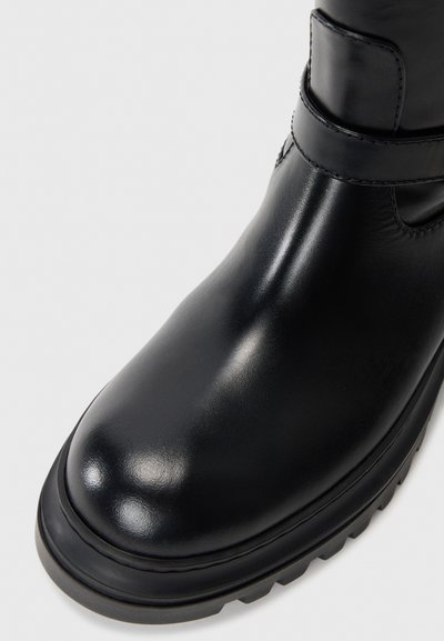 Black leather ankle boot with a rounded toe, reinforced stitching, and rubber outsole. Features a unique strap detail across the upper.