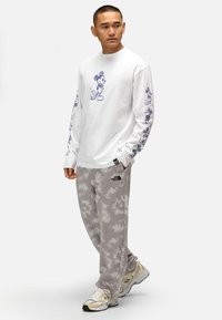 Camicia bianca a maniche lunghe con stampa blu di Topolino sul davanti e sulle maniche, abbinata a pantaloni della tuta grigi tie-dye e sneakers beige.