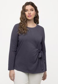 Marineblaues Langarmoberteil mit Rundhalsausschnitt, das über ein seitliches Knotendetail und einen weichen Stoff verfügt. Minimalistisches Design, lässige Passform, kombiniert mit weißen Hosen.
