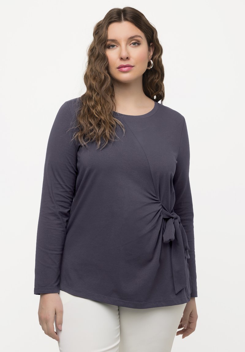 Marineblaues Langarmoberteil mit Rundhalsausschnitt, das über ein seitliches Knotendetail und einen weichen Stoff verfügt. Minimalistisches Design, lässige Passform, kombiniert mit weißen Hosen.
