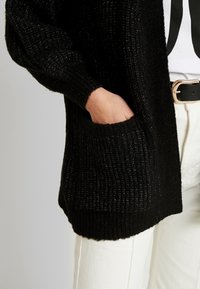 Schwarzer Strickcardigan mit strukturiertem Muster, ausgestattet mit einer Fronttasche und gerippten Bündchen. Kombiniert mit hellen Hosen und einem schwarzen Gürtel.