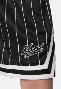 Černé sportovní šortky s bílými vertikálními pruhy, vyrobené z prodyšné tkaniny, s bílým lemem a vyšitým logem s nápisem "est. 1989."