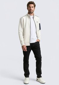 Witte jas met ritssluiting en gestructureerd patroon, marineblauwe accenten, en zijzakken, gecombineerd met een wit T-shirt, zwarte spijkerbroek en witte sneakers.