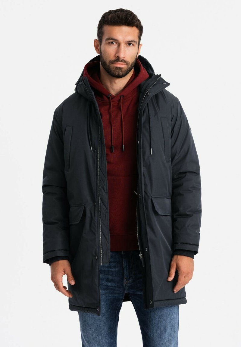 Dunkelraffinierte Daunenjacke mit Kapuze, die zwei vordere Taschen, einen Reißverschluss und Kordelzüge hat. Über einem burgunderfarbenen Hoodie und blauen Jeans getragen.