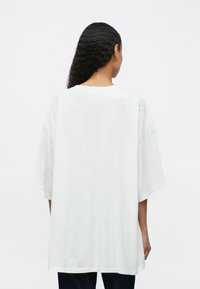 Άτομο με μακριά σγουρά μαλλιά που φοράει ένα ολόσωμο λευκό T-shirt και σκούρους παντελόνες, γυρισμένο μακριά με φόντο μια απλή λευκή επιφάνεια.