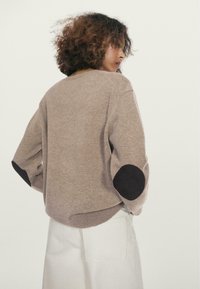 Lysebrun strikket sweater med løs pasform, med sorte albuebeskyttere og rundet kant, kombineret med hvide bukser.