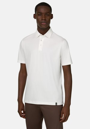 Jeune homme portant un polo blanc uni à manches courtes et un pantalon marron, debout devant un fond clair uni.