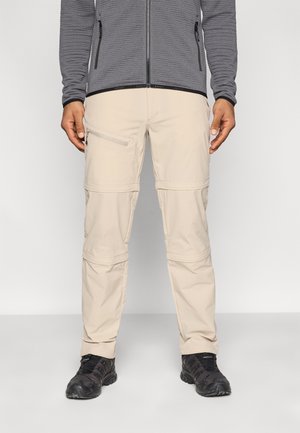 Μπεζ παντελόνι cargo με τσέπες με φερμουάρ, με λεία υφή και ρυθμιζόμενες μανσέτες, σε συνδυασμό με γκρι υφασμάτινο μπουφάν με φερμουάρ.