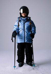 Dijete s pletenicama nosi plavi skijaški kaput, crne hlače, kacigu, naočale i rukavice, držeći skijaške štapove dok stoji na snijegu.