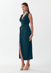 Femme debout portant une robe portefeuille sans manches bleu sarcelle foncé avec un col en V profond, une fente sur le côté, des boucles d'oreilles dorées, un bracelet et des sandales à talons hauts à brides.