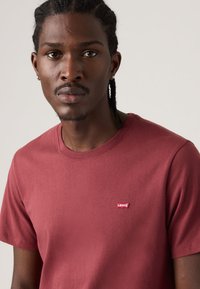 Mladý černoch s copánky, oblečený v jednoduchém červeném tričku Levi’s, stojí čelem vpřed na neutrálním pozadí.