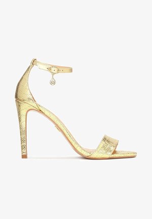 Sandale dorée à talons hauts avec une finition texturée, une fine sangle à la cheville et une bande à l’avant. Elle dispose d'un petit charm sur la sangle pour plus de détail.