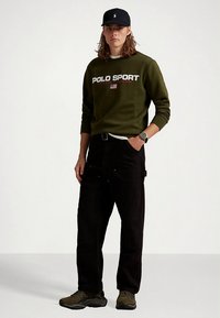 Φούτερ σε ελιά πράσινο με το κείμενο "POLO SPORT", μαύρο παντελόνι cargo με τσέπες και σκούρα πράσινα αθλητικά παπούτσια. Διακριτό μαύρο καπέλο και ρολόι χειρός.