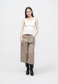 Witte tanktop, beige broek met wijde pijpen en een riem, en zwarte, puntige enkellaarsjes. Model staat tegen een effen achtergrond.