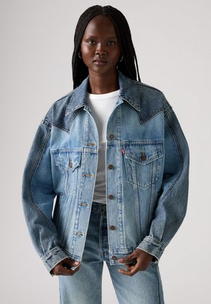 Giacca in denim azzurro chiaro con collare strutturato, due tasche sul petto, dettagli cuciti e una vestibilità rilassata, abbinata a un top bianco.