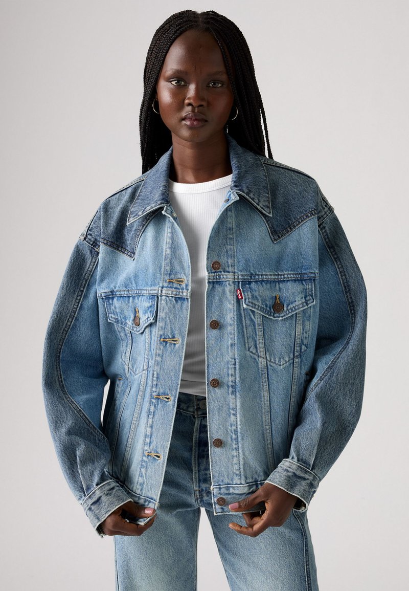 Veste en denim bleu clair avec un col structuré, deux poches poitrine, des détails cousus et une coupe décontractée, associée à un haut blanc.
