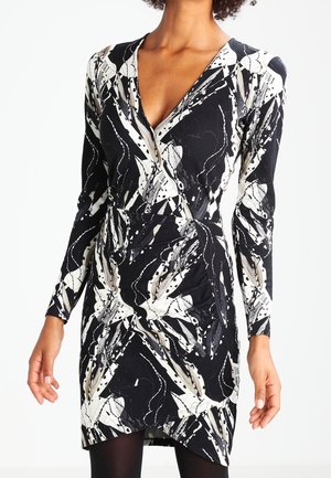 Nainen pukeutunut mustavalkoiseen abstraktiin kuviolliseen pitkähihnaiseen mekkoon, jossa on V-pääntie ja vyötäröllä kerätty yksityiskohta.