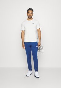 Hvid T-shirt med korte ærmer og sort logo, parret med blå joggebukser med hvide sidelommer og hvide sneakers. Holder et sjippetov.