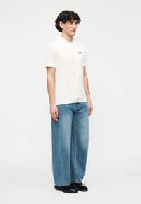 Polo branca de manga curta com um logótipo bordado no peito, combinada com jeans azuis de perna larga e sapatos pretos.