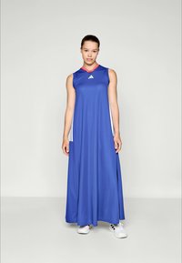 Jeune femme portant une longue robe de sport sans manches bleu avec un col rouge et des baskets blanches, se tenant devant un fond clair uniforme.