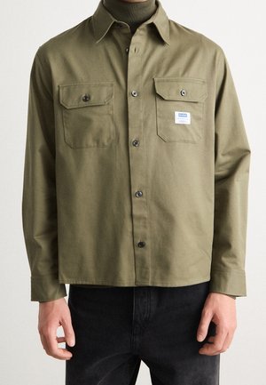 Mężczyzna ubrany w oliwkowo-zieloną koszulę na guziki z dwoma zapinanymi na guziki kieszeniami na piersiach, z których jedna ma małą białą metkę Hugo, w połączeniu z czarnymi spodniami.