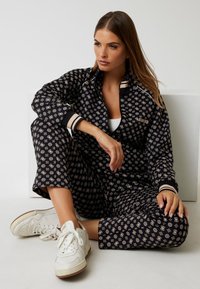 Tuta nera con motivo, presentante disegni beige, con polsini e colletto a righe. Abbinato a sneakers bianche su una superficie chiara.
