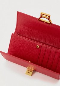 Červená texturovaná kožená peněženka otevřená, ukazující sloty na karty a zlatou sponu s názvem značky na světlém pozadí.