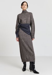 Robe en tricot grise avec un col montant et des manches longues, dotée d'une ceinture en tissu bleu marine. Le mannequin porte des bottes aiguilles noires brillantes.