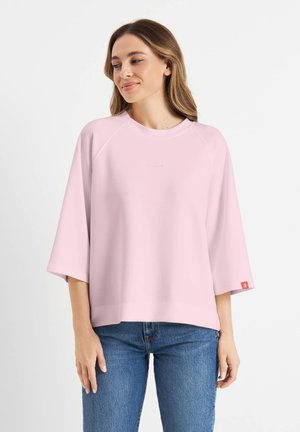 Vrouw in een lichtroze oversized trui met driekwartmouwen en een blauwe spijkerbroek, staand tegen een witte achtergrond.