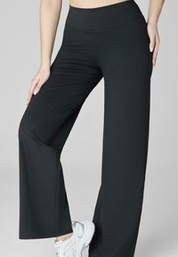 Czarne spodnie z szerokimi nogawkami, wykonane z elastycznego materiału, z wysokim stanem i gładką teksturą, w połączeniu z białymi butami sportowymi.