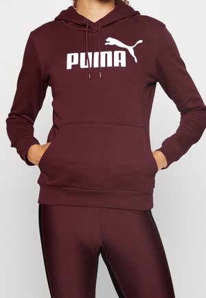 Osoba nosząca bordową bluzę z kapturem Puma z kieszenią kangura i pasujące legginsy na jednolitym tle.