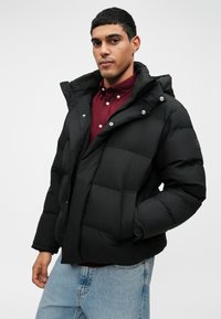 Juodas pūkinis švarkas su kapišonu, turintis siuvinėto dizaino, priekinėmis kišenėmis ir spaudėmis užtrauktukais. Dėvimas virš raudonai vyno spalvos marškinių su mygtukais.