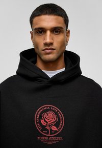 Zwarte hoodie met een rode roos graphic en tekst in een cirkelontwerp. Stof lijkt zacht en gestructureerd; inclusief een trekkoordcapuchon.