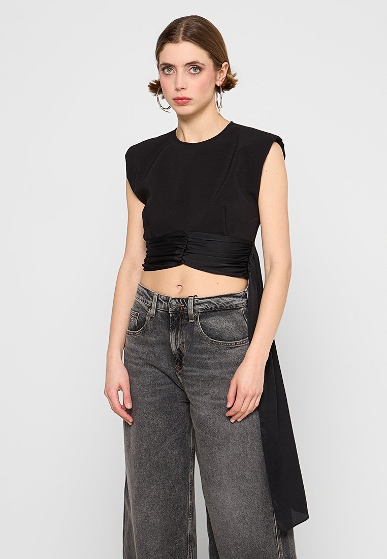 Schwarzes, cropped Top mit strukturierten Schultern und gerafftem Bund, ausgestattet mit einem langen Satinband; kombiniert mit hochtaillierten, dunklen Jeans.