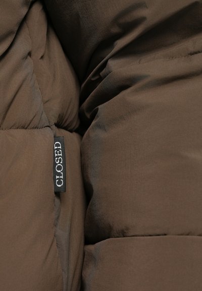 Giacca a vento marrone in tessuto morbido, con sezioni trapuntate e un'etichetta nera con la scritta "CLOSED" cucita nella cucitura.