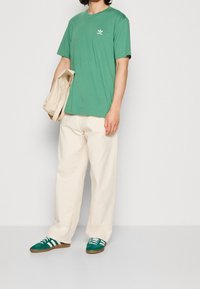 Grön bomullströja med vitt logotyp, krämfärgade vida byxor och gröna skor med vita ränder. Slät textur, avslappnad passform.