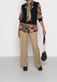Czarny bezrękawnik na kolorowej bluzce w nadruk paisley, beżowe szerokie spodnie i czarne buty, z małą czarną torebką.