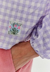 Fioletowa tkanina w kratkę z kwiatową łataną łatą z tulipanami i napisem "TULIP." Częściowo widoczny jest różowy materiał sztruksowy pod spodem.