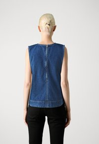 Haut en denim bleu sans manches avec un col montant, fermeture éclair au dos et surpiqûres contrastées ; porté avec un pantalon noir. Design simple et structuré.