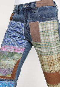 Jeans de mezclilla con un diseño de patchwork que presenta patrones textiles coloridos, acentos de ante marrón y diversas texturas de tela en varios colores.