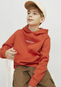 Felpa arancione con tasca a marsupio e logo in testo bianco, abbinata a un cappellino beige e pantaloni khaki, seduto su una sedia bianca.