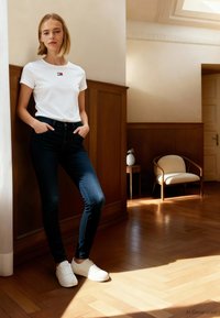 T-shirt branca de algodão com um pequeno logótipo bordado, jeans skinny azul escuro e sapatilhas brancas, com um cenário de interior em madeira.