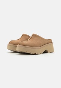 Zoccoli in suede color tan con una piattaforma elevata, punta arrotondata e dettagli cuciti lungo il bordo, dotati di una suola in gomma testurizzata per una migliore aderenza.