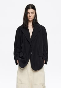 Blazer oversize nero con rever a punta, chiusura a un bottone e due tasche frontali. Realizzato in tessuto morbido con una texture liscia.