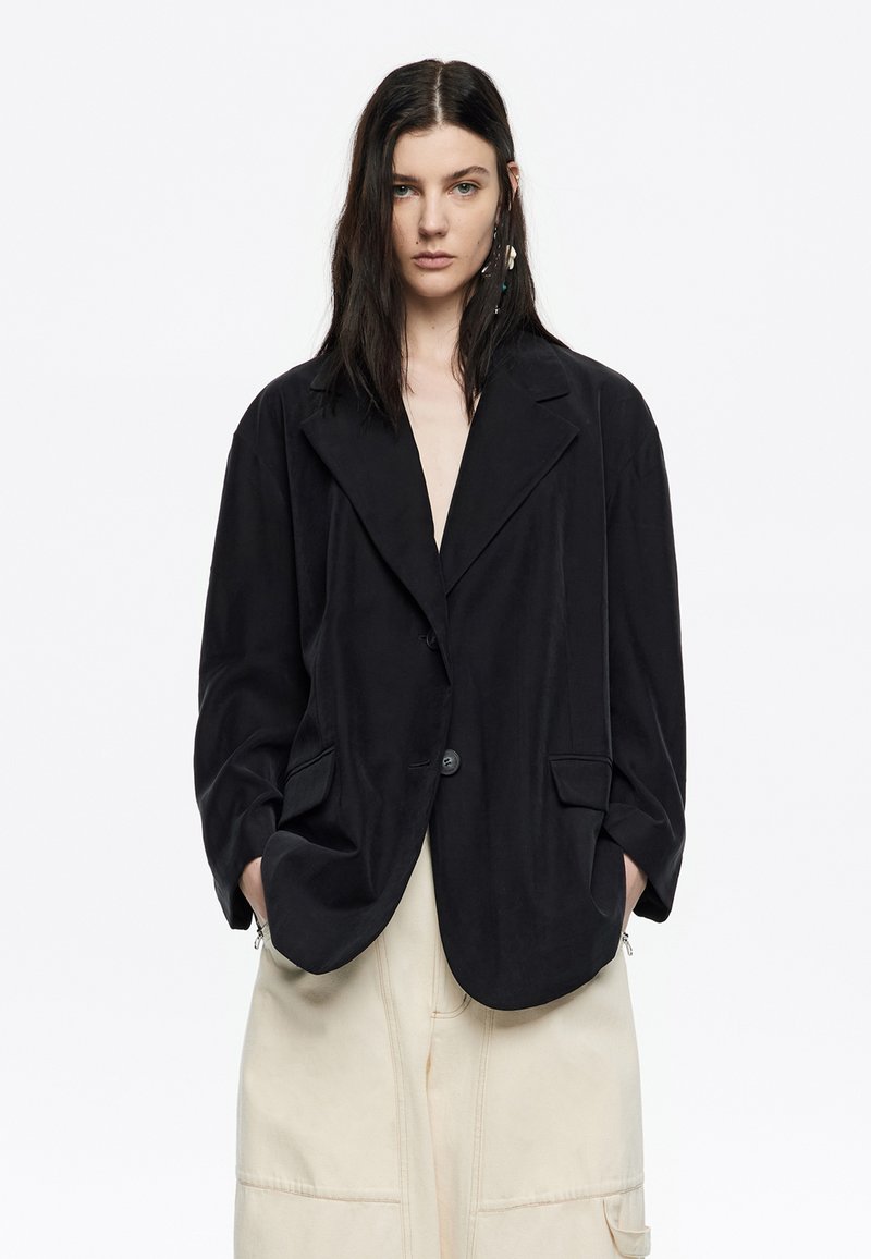 Blazer oversize nero con rever a punta, chiusura a un bottone e due tasche frontali. Realizzato in tessuto morbido con una texture liscia.
