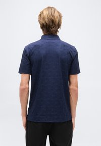 Homme aux cheveux blonds courts, vu de dos, portant une chemise à manches courtes bleu marine à motifs et un pantalon noir devant un fond uni.