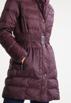 Mujer con abrigo de invierno acolchado de color púrpura oscuro, de longitud media y con cinturón, con bolsillos con cremallera y cierre de botones, de pie con una mano en el bolsillo.