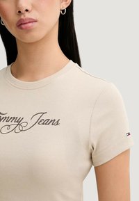Béžové žebrované tričko s krátkými rukávy, s logem "Tommy Jeans" v hnědé barvě a malým detailem vlajky v červené, bílé a modré barvě na rukávu.